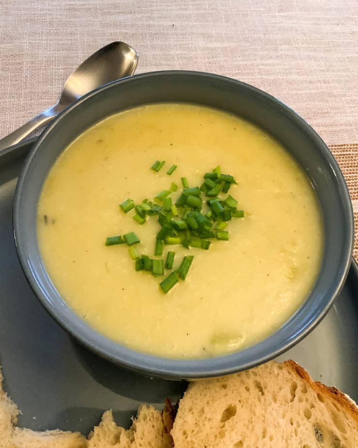 Creamy sou[ with chopped green chives on top in a blue bowl with a silver soup spoon on the top left side and fresh bread to side of bowl