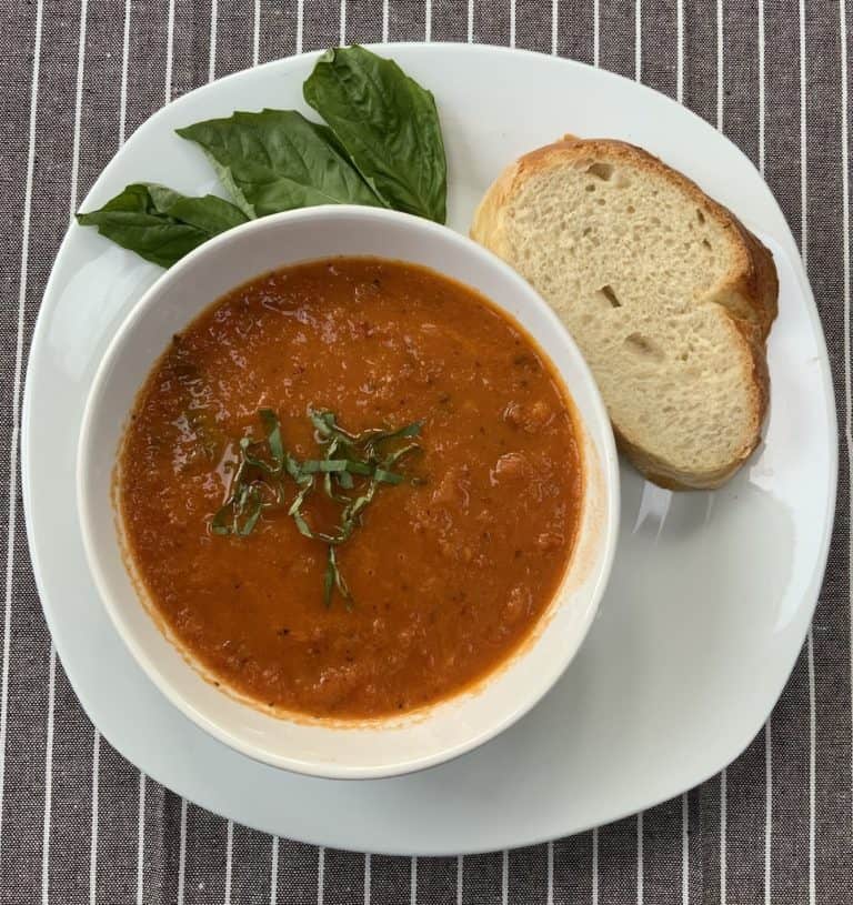 Roasted tomato soup in a white bowl garnished with sliced fresh green basil. Soup bowl in on a white plate that has a sliced of fresh white bread and fresh basil leaves.