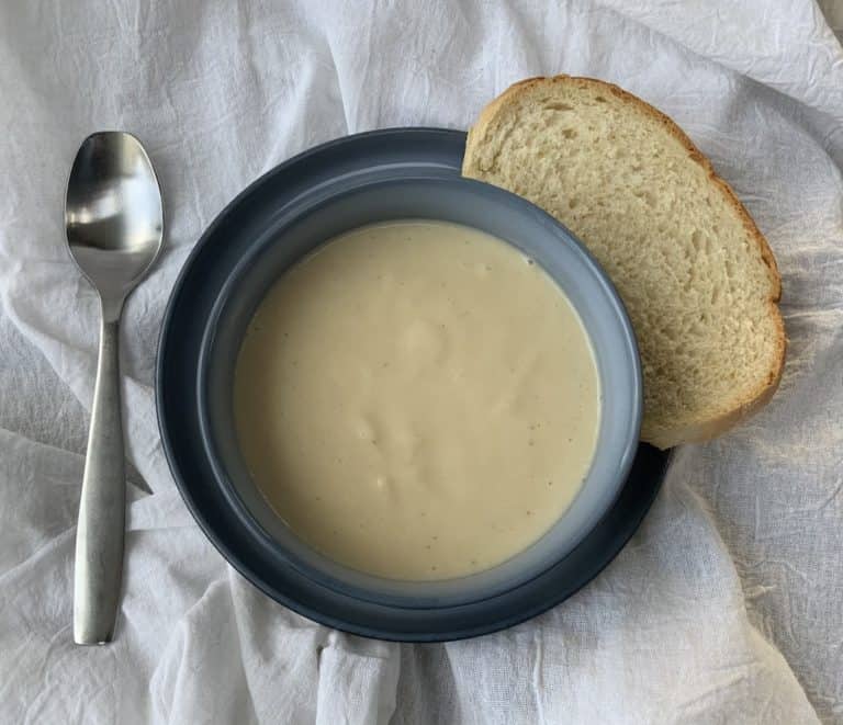 pureed white soup in a blue bowl on a blue plate with a slice of fresh bread on side of plate. Plate is on a white linen surface with a silver soup spoon to the left of the soup.