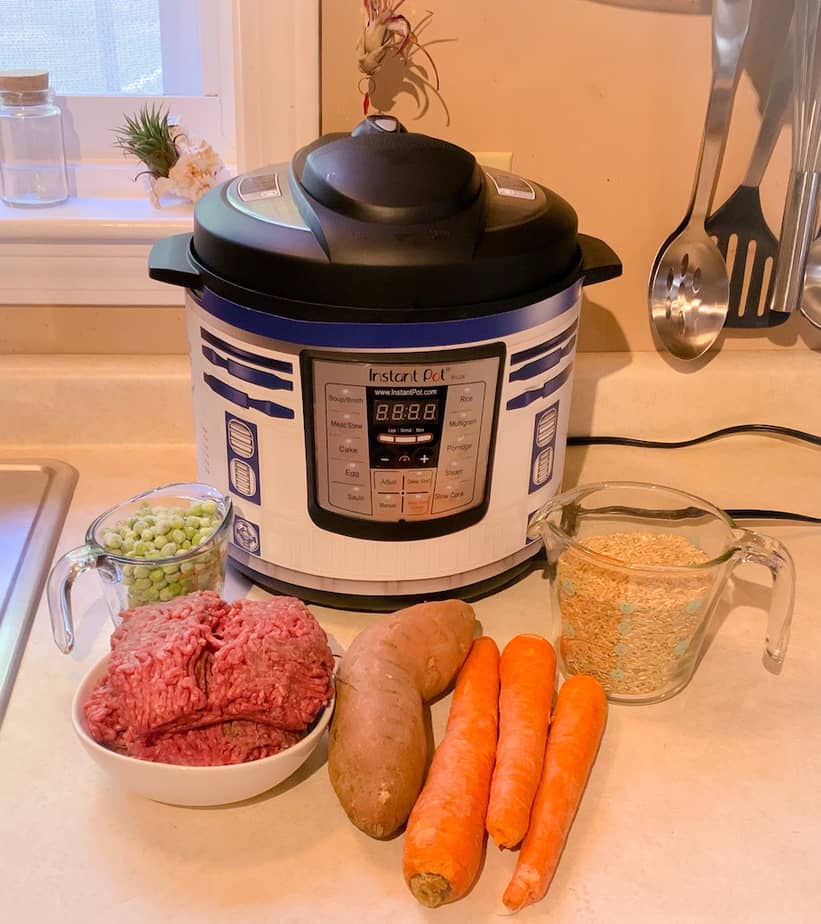 Instant pot with measuring glass of peas, bowl of raw Hamburg, sweet potato, three carrots, measuring glass of brown rice
