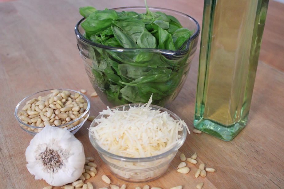 three clear glass bowls, one filled with fresh basil leaves, one filled with pine nuts, another filled with fresh Parmesan, bulb of garlic and glass jar of olive oil. 