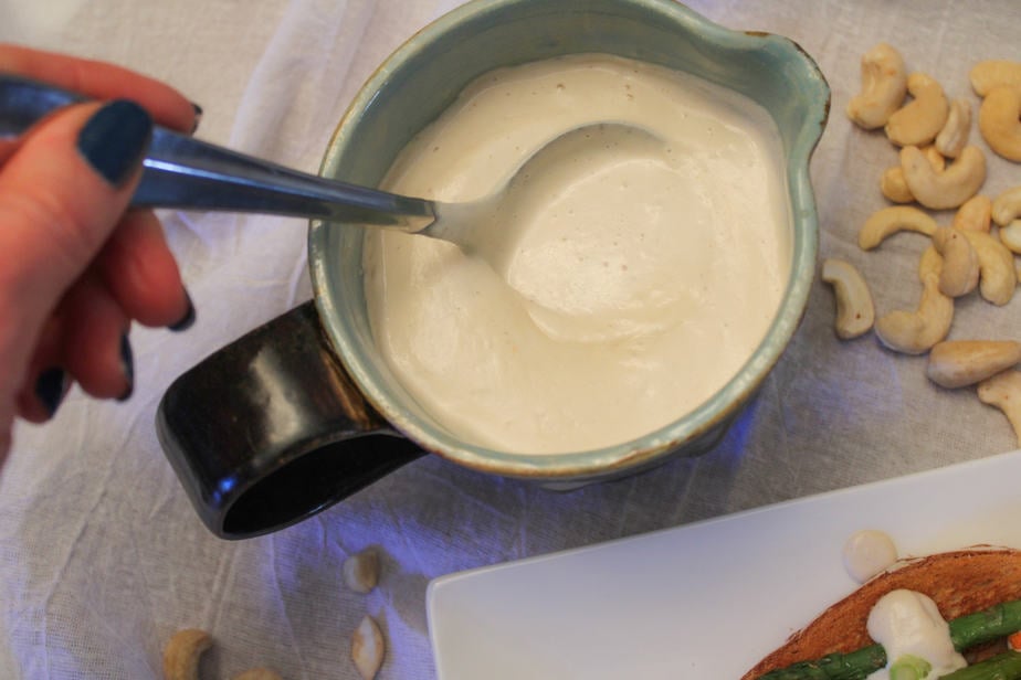 Cashew sauce in a gravy bowl with whole cashews to the upper right and a white plate with a corner of toast, vegetable and cashew sauce on it.