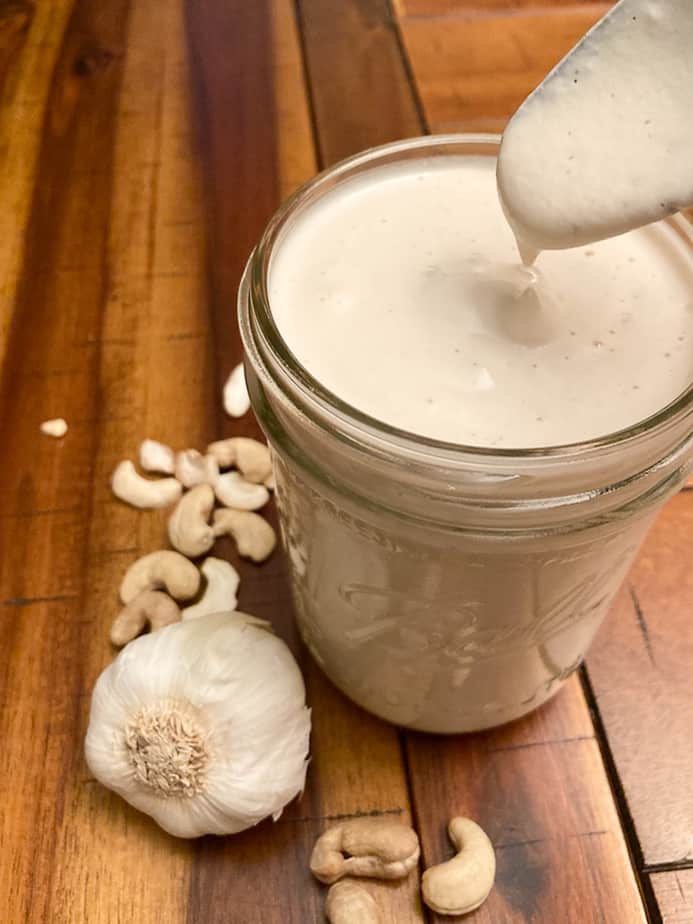 Roasted garlic cashew sauce in a canning jar, with pouring sauce in glass. Fresh garlic bulb and cashews in surface next to sauce. 