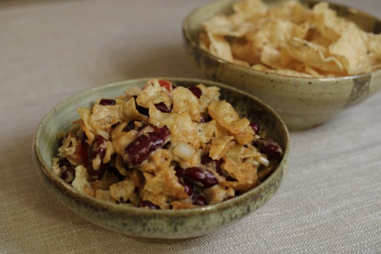 Red kidney beans with a sauce, cheese and crushed tortilla chips on top in a ceramic bowl with another ceramic bowl filled with tortilla chips in it.