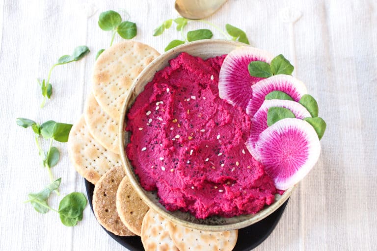 red beet hummus in a bowl with sliced watermelon radish and green leaves decorating the bowl and background on a cream colored linen. crackers surround the left side of plate under bowl with hummus.