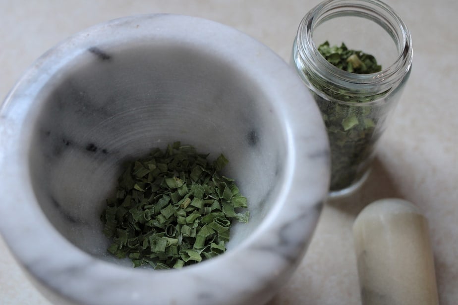 Marble mortar with dried green chives, marble pestle to the side and clear glass jar of dried herbs at top of photo