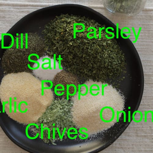 Ingredient spices on a black plate with labels reading top left clockwise - parsley, pepper, onion, chives, garlic, salt, dill.