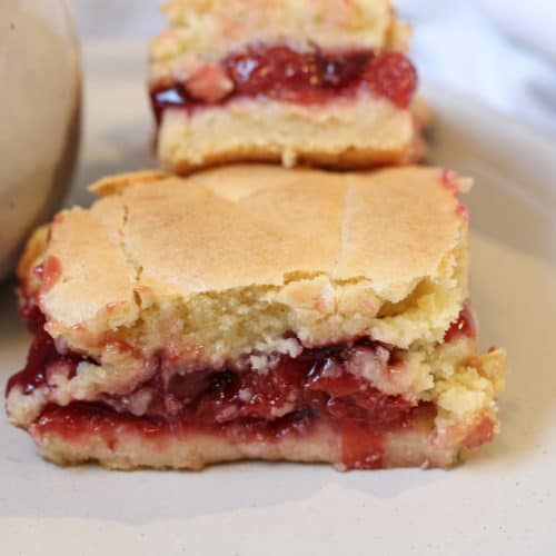 A square cherry dessert with cake-like bottom and top, cherry filling in the center of dessert. Another square stacked to the back of first cherry dessert. Both on a white plate.
