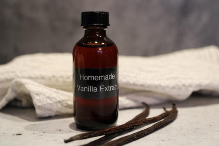 Homemade Vanilla Extract in an amber colored bottle, labeled Homemade Vanilla Extract. Whole beans in front of bottle with white towel behind bottle.