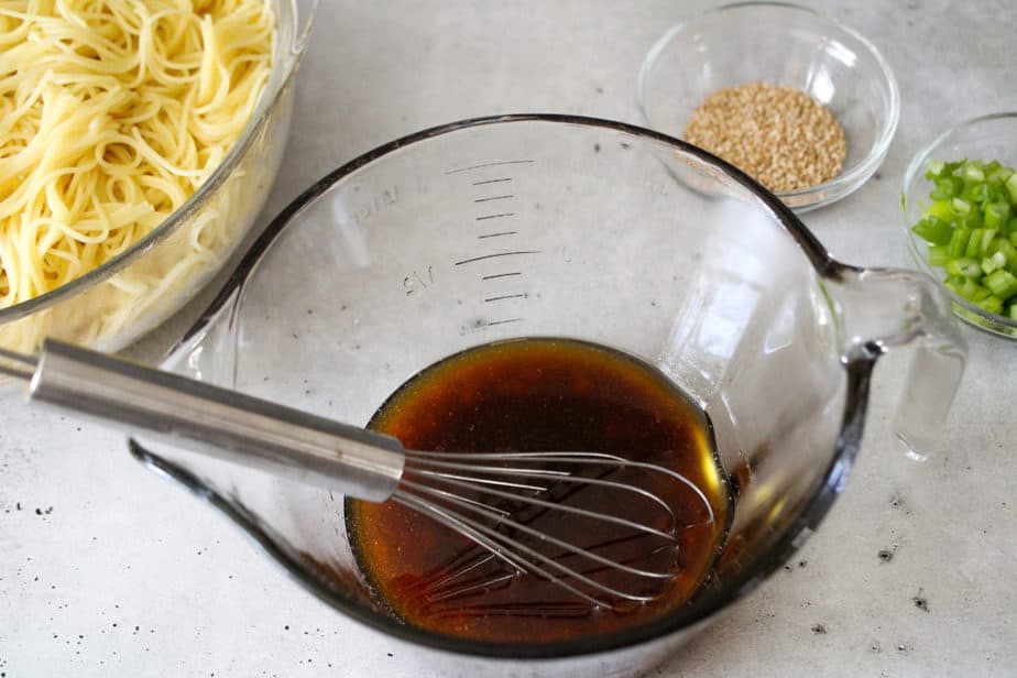 Sesame noodle sauce in clear glass bowl with cooked noodles in glass bowl top left and small glass bowl of sesame seeds and small glass bowl of chopped green onions to the top right.