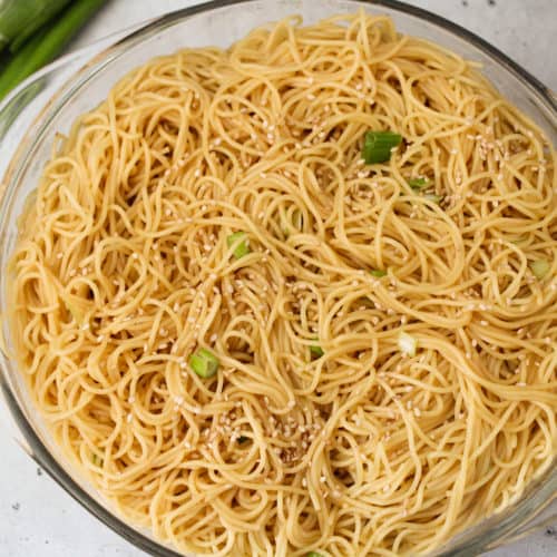 Angel hair noodles with sesame sauce and sesame seeds in a glass bowl with green onions and small glass bowl of sesame seeds in the background.