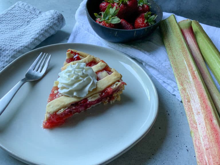 Slice of strawberry rhubarb pie on a plate. Wooden bowl of fresh strawberries and stalks of rhubarb in the background.