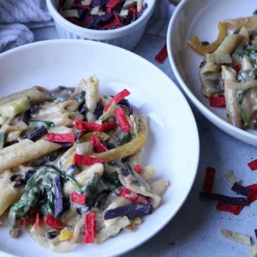 Pasta with creamy sauce with vegetables and colored tortilla strips in a white bowl.
