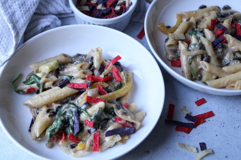 Pasta with creamy sauce with vegetables and colored tortilla strips in a white bowl.