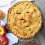 Homemade apple pie with apples and cinnamon sticks on surface around the baked pie.