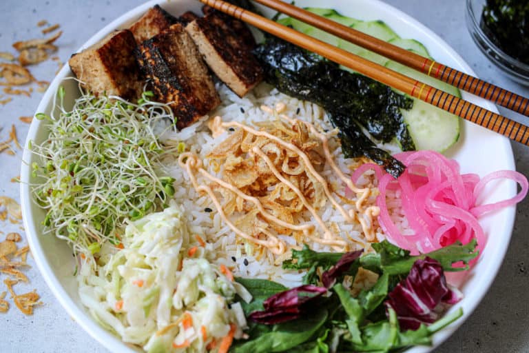 Tofu poke bowl with rice, tofu, sliced nori, pickled red onions, lettuce, Cole slaw, bean sprouts, crispy onions in center drizzled with spicy mayo.