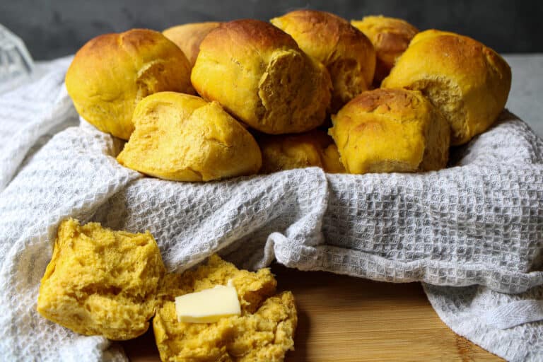 Pumpkin dinner rolls in cloth covered bowl with one roll on surface in front of bowl cut in half with butter on top.