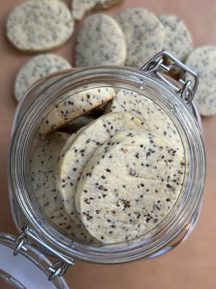 Round thick cookies with chia seeds in a clear glass jar with additional cookies on surface surrounding jar.