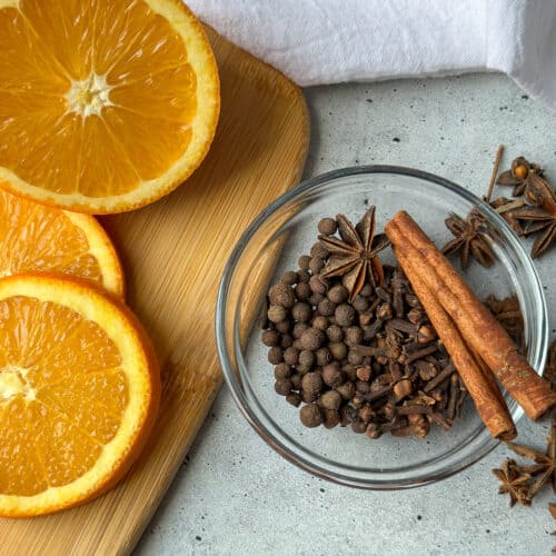 Mulling spices in a small clear glass bowl with fresh orange slices to the left.