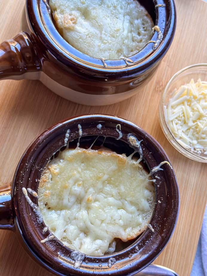 Vegetarian French Onion Soup in two brown ceramic tureens with broiled French bread and cheese on top.