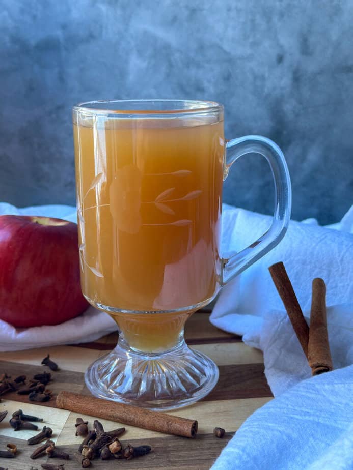 Hot mulled cider in a clear glass mug with spices on the surface.