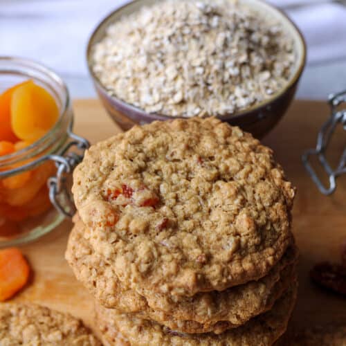 Oatmeal apricot cookies stacked with quick oats, apricots and pecans in the background.