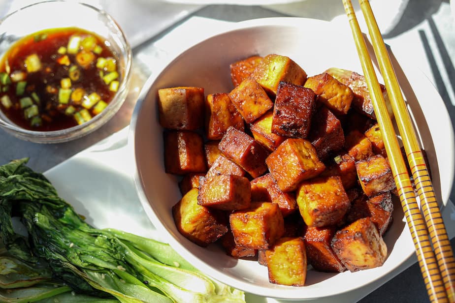 Crispy air fried tofu, cubed, in a white bowl with dipping sauce in small bowl at top left, and cooked bok choy to bottom left. 