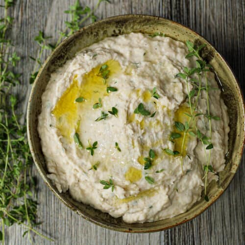 White bean and fresh thyme dip in a brown ceramic bowl with fresh thyme leaves sprinkled on top.