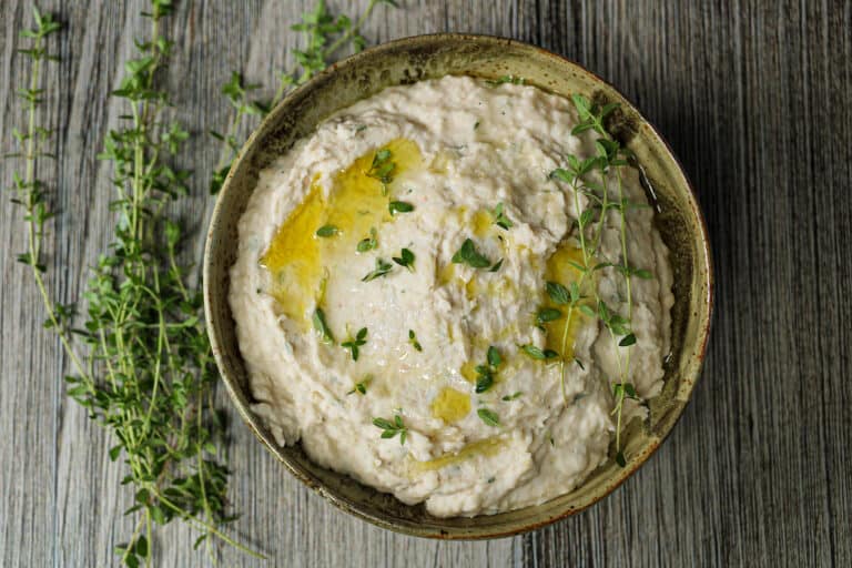 White bean and fresh thyme dip in a brown ceramic bowl with fresh thyme leaves sprinkled on top.