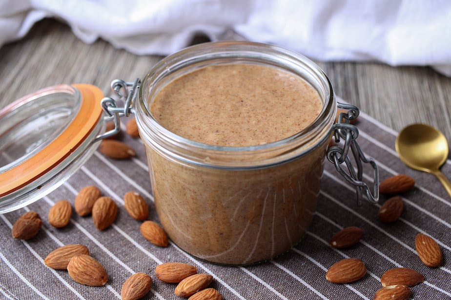 Creamy almond butter in a glass jar with whole almonds and gold spoon on surface surrounding the jar.