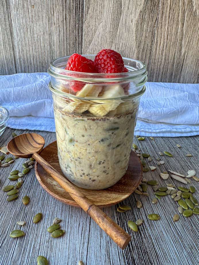 Glass mason jar with overnight oat mixture and sliced bananas and fresh raspberries on top. Jar is on a wooden plate with a wooden spoon to the left side.