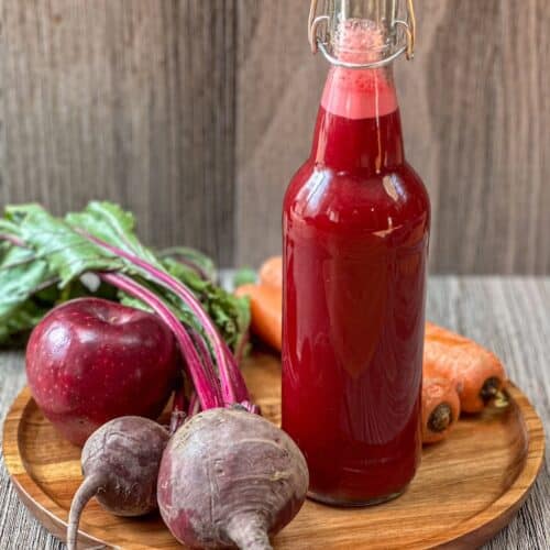 Bright red beet and carrot juice in a glass bottle with whole beets, apple and carrots on surface.