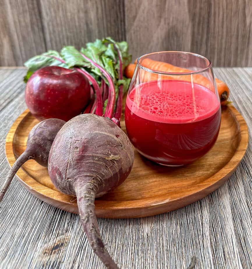 Glass of beet and carrot juice which is surrounded by fresh beets, carrots and apple.