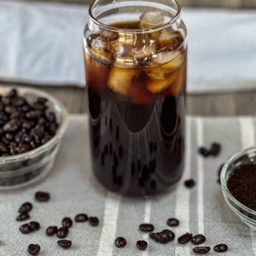 Clear glass filled with cold brew coffee over ice. Small bowls of coffee grounds and beans to each side and coffee beans scattered on surface.