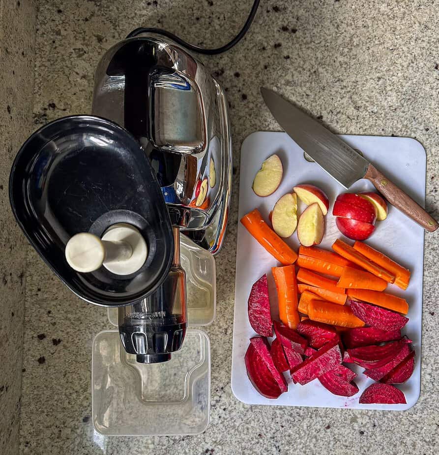 Above shot of high powered juicer with sliced beets, carrots, and apple on white cutting board with kitchen knife to the side.