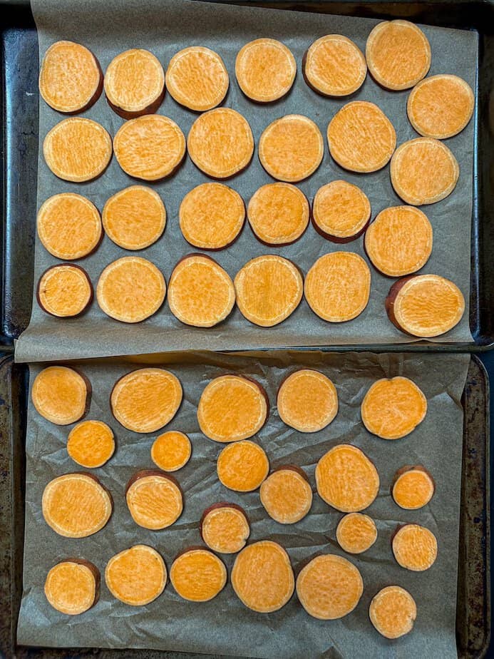 Single layer of sliced sweet potatoes arranged on two baking sheets.