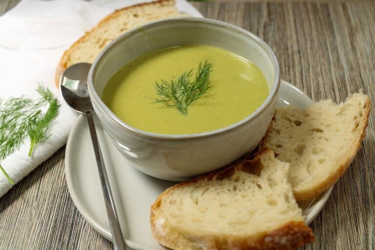 Creamy green asparagus soup with a sprig of dill on top in a ceramic bowl with fresh bread to side of bowl.