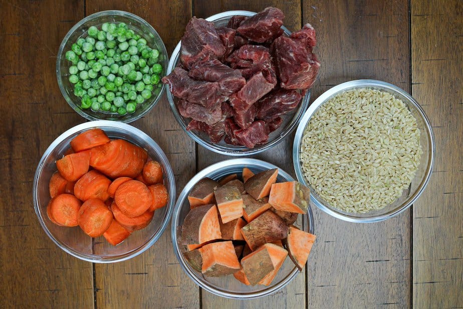 Ingredients for stew beef dog food, counterclockwise at top, bowl of stew beef, brown rice, cubed sweet potatoes, large slices of carrots, and bowl of peas.