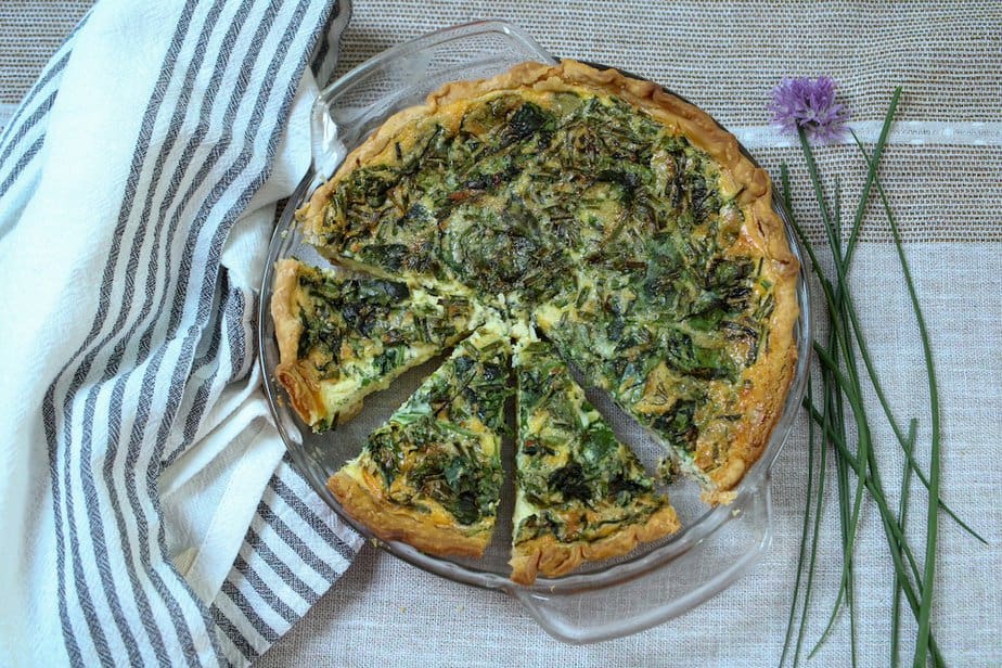 Baked quiche with three slices cut and chives and a chive blossom to side of pie plate.