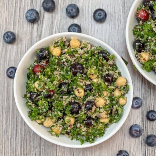 Kale Salad with quinoa, blueberries, chick peas in a white bowl with fresh blueberries on the surface surrounding the bowl.