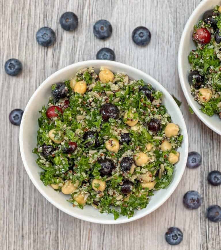 Kale Salad with quinoa, blueberries, chick peas in a white bowl with fresh blueberries on the surface surrounding the bowl.