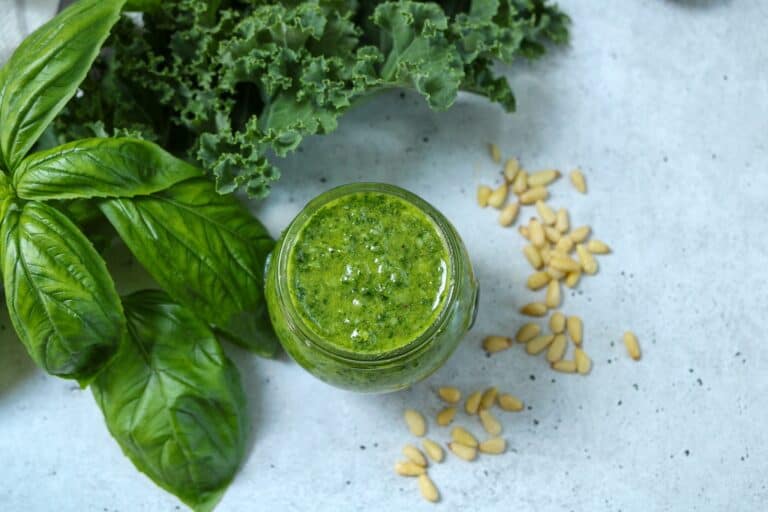 Kale and basil pesto in a glass jar with pine nuts, basil leaves and kale on surface surrounding the jar.