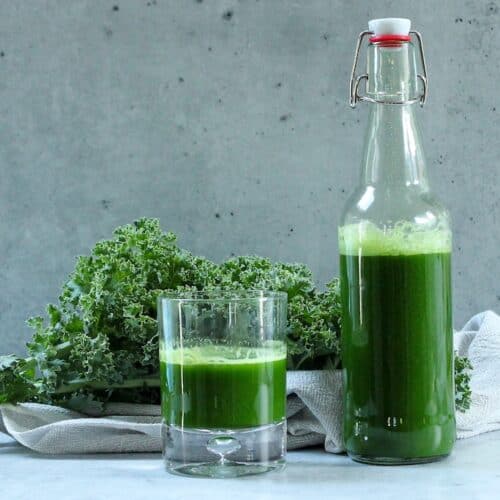 Kale and cucumber juice in small glass and a large bottle with kale in background.