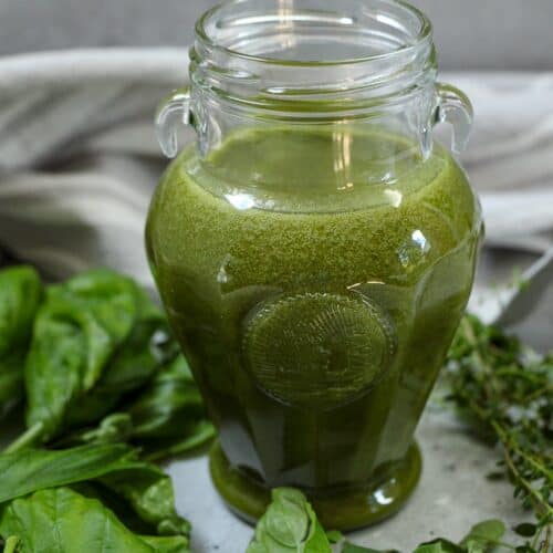 Fresh green herb oil in a glass jar with fresh herbs around the surface of the jar.