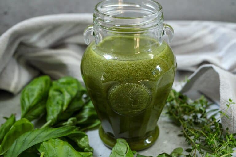 Fresh green herb oil in a glass jar with fresh herbs around the surface of the jar.