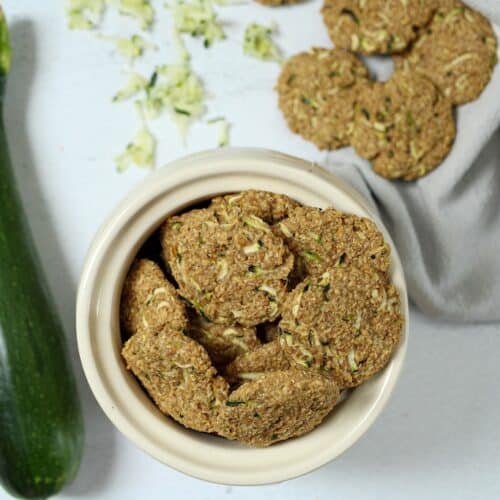 Round zucchini dog treats in a ceramic container with whole zucchini and shredded zucchini on surface.