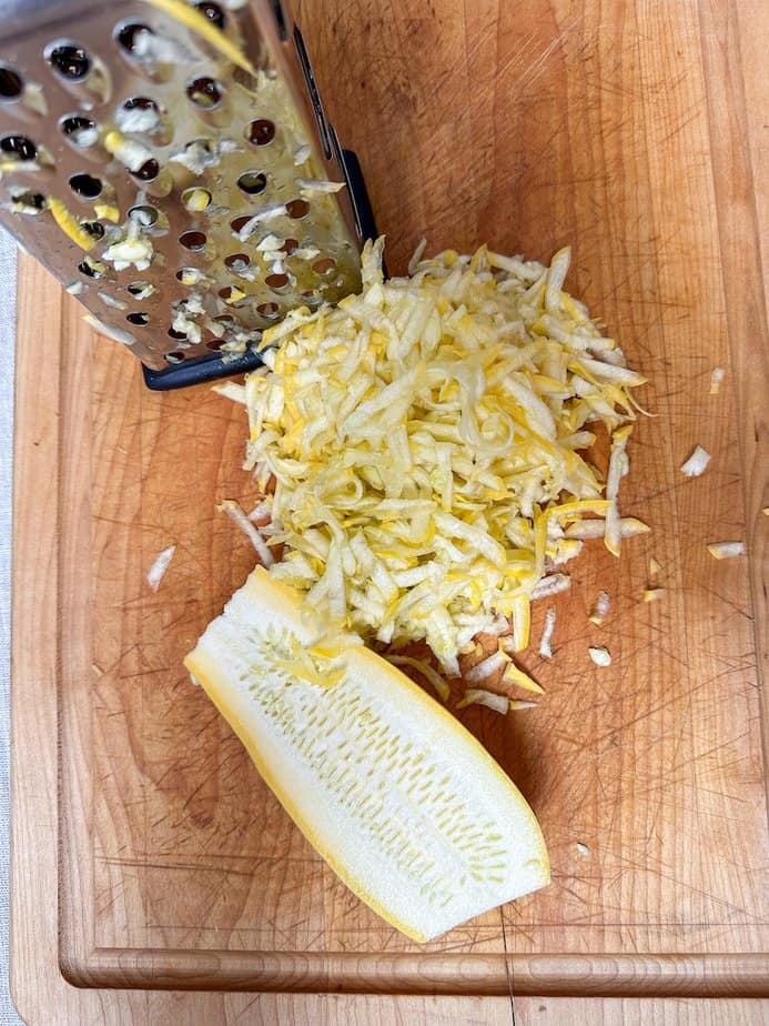 Shredded summer squash in center with mix grater at top left and halved summer squash at the bottom of the photo. 