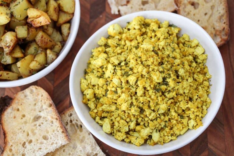 Scrambled tofu in a white bowl with toast and skillet potatoes in a bowl to the side.