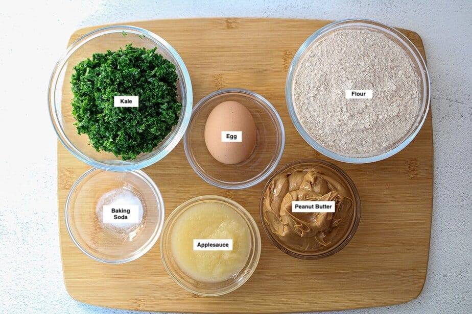 Individual bowls filled with dog cookies ingredients which include: minced kale, one egg, flour, creamy peanut butter, applesauce and baking soda.