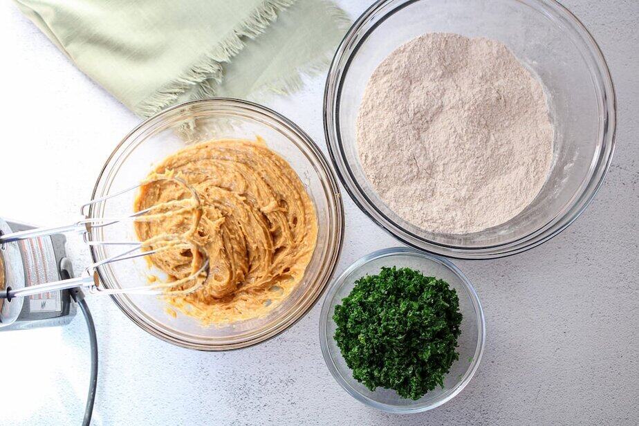 Three clear glass bowls, one with dry flour ingredients, one with wet batter, and the other with minced green kale.